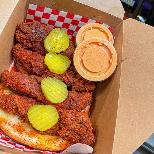 Nashville Coop's hot fried chicken tenders with pickles and dipping sauce in a takeout box.