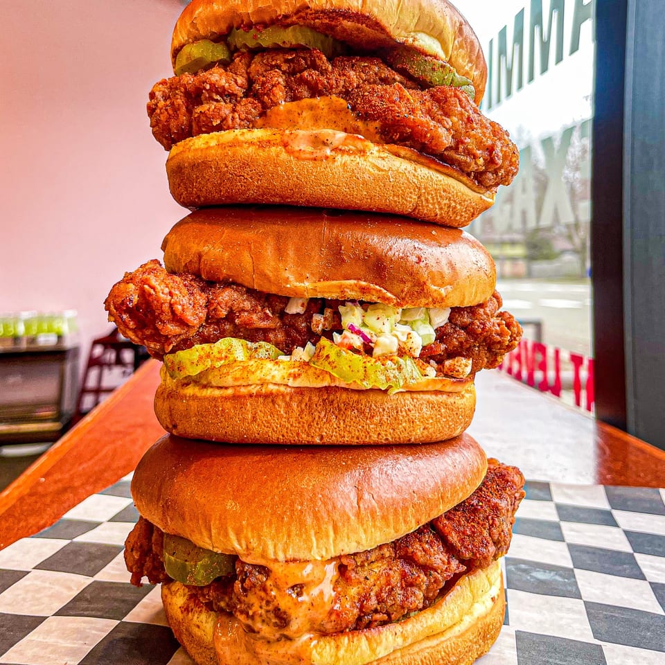 A towering stack of three Nashville Coop fried chicken sandwiches with pickles and slaw.
