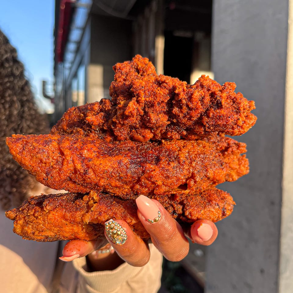 A hand with manicured nails holding three crispy, red Nashville Coop fried chicken tenders outdoors.